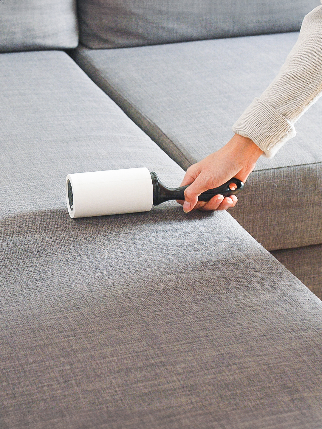 A hand holding a lint roller is cleaning the fabric of a gray sectional sofa.