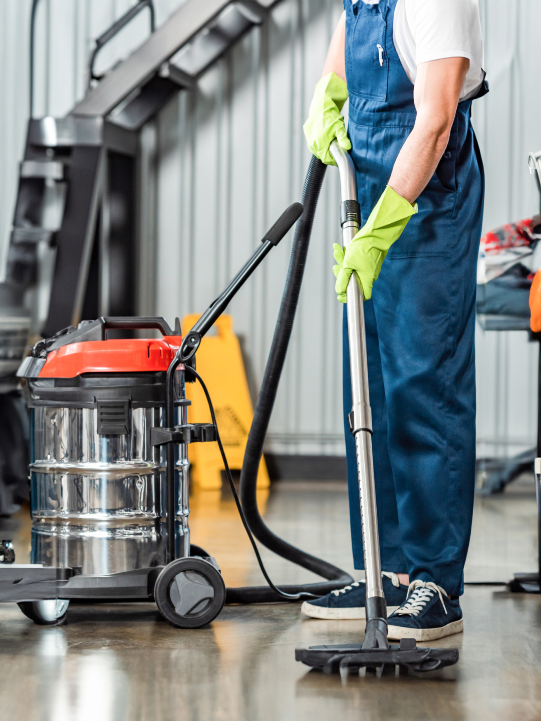 A person wearing green gloves and blue overalls is using a vacuum cleaner. The vacuum is on wheels and has a shiny metal body. The background features industrial shelving and cleaning supplies.