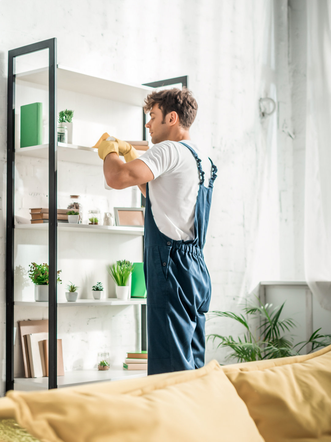 A person in a white t-shirt and blue overalls is cleaning a shelf with potted plants in a bright, modern living space.